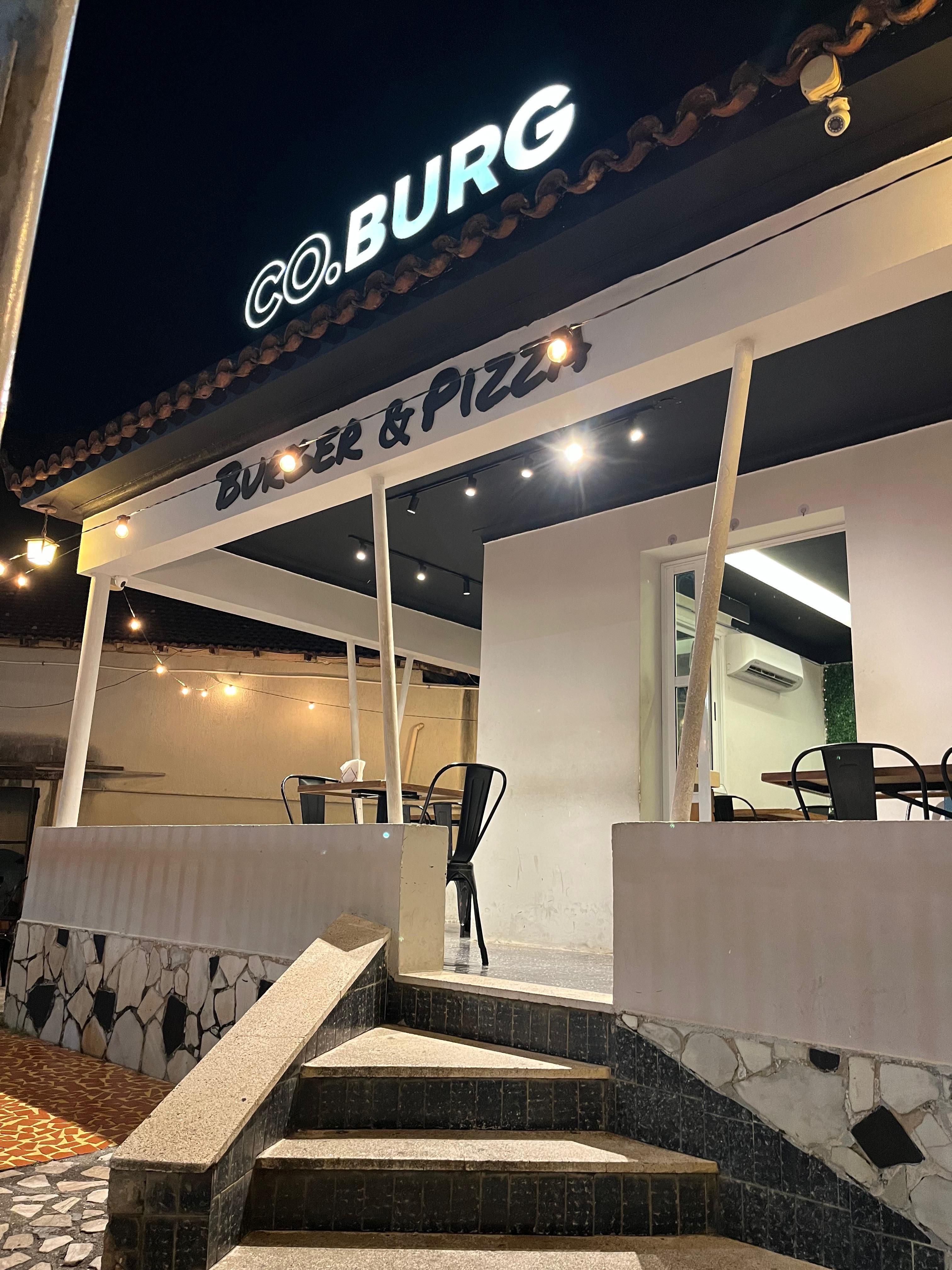 Fachada da Co.Burg à noite, com o letreiro aceso e a varanda com mesas